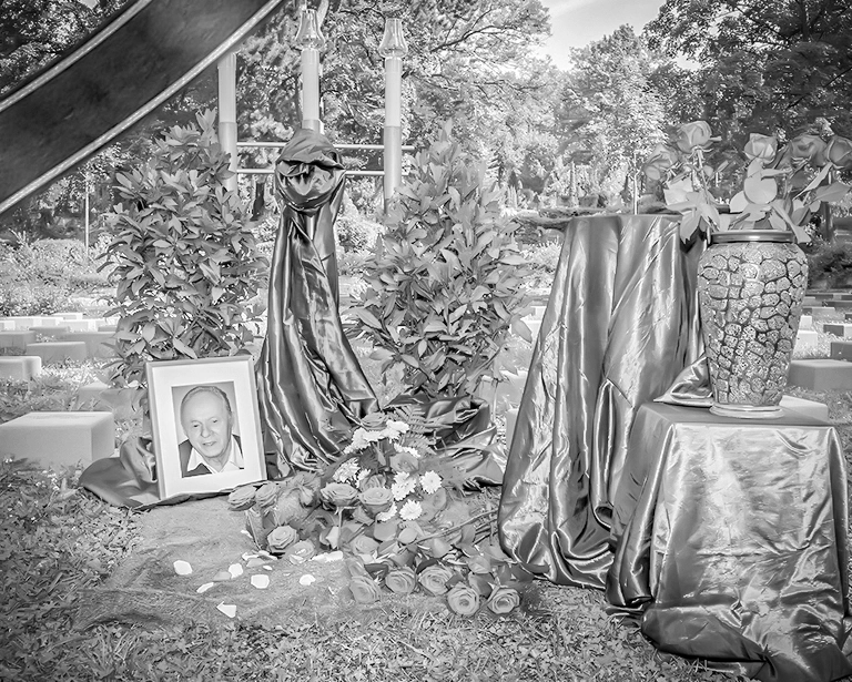 Eine Urne mit einem gerahmten Porträtfoto eines verstorbenen Mannes inmitten von Blumen und Tüchern bei einer Urnenbestattung auf einem Friedhof, symbolisierend das Gedenken an geliebte Menschen. Das Schwarz-Weiß-Foto unterstreicht die feierliche Atmosphäre.